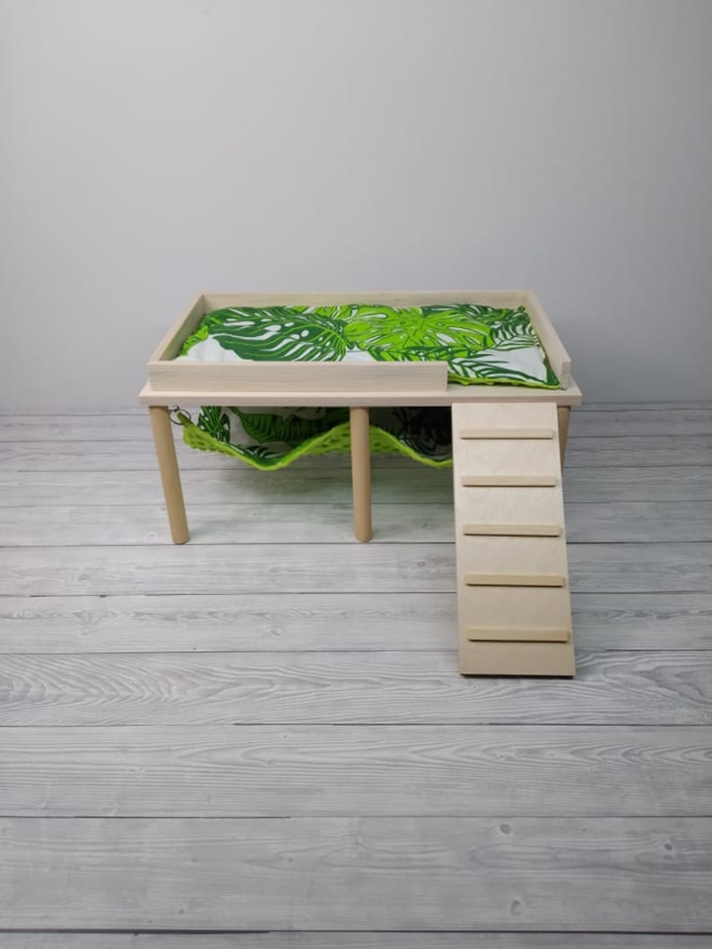 May include: A light wood pet bed with a green and white leaf-patterned cushion. The bed has a built-in ladder and a hammock underneath. The bed is designed for small pets.