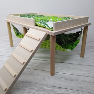 May include: A wooden pet habitat with a raised platform, a ramp, and two hammocks. The platform has a green and white tropical leaf-patterned cushion. The hammocks are green with a similar leaf pattern. The habitat is made of light-colored wood.