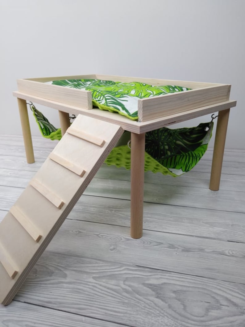 May include: A wooden pet habitat with a raised platform, a ramp, and two hammocks. The platform has a green and white tropical leaf-patterned cushion. The hammocks are green with a similar leaf pattern. The habitat is made of light-colored wood.
