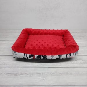 K&ouml;nnte beinhalten: Ein rotes und wei&szlig;es Haustierbett mit einem gemusterten Stoff, der schwarz-wei&szlig;e Illustrationen von Frauen zeigt. Das Bett hat einen erh&ouml;hten Rand und eine weiche, rote, gepunktete Stoffauskleidung.