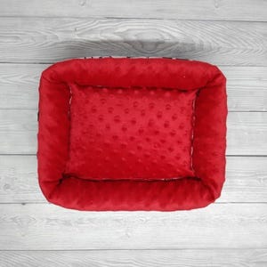 K&ouml;nnte beinhalten: Rotes Haustierbett mit erh&ouml;htem Rand und weicher, strukturierter Auskleidung. Das Bett ist mit einem pl&uuml;schigen, roten Kissen gef&uuml;llt.