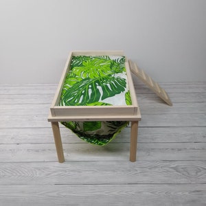 May include: A small wooden pet bed with a green and white tropical leaf print fabric. The bed has a rectangular frame, four legs, and a hammock-style bottom. A small wooden ladder is leaning against the side.