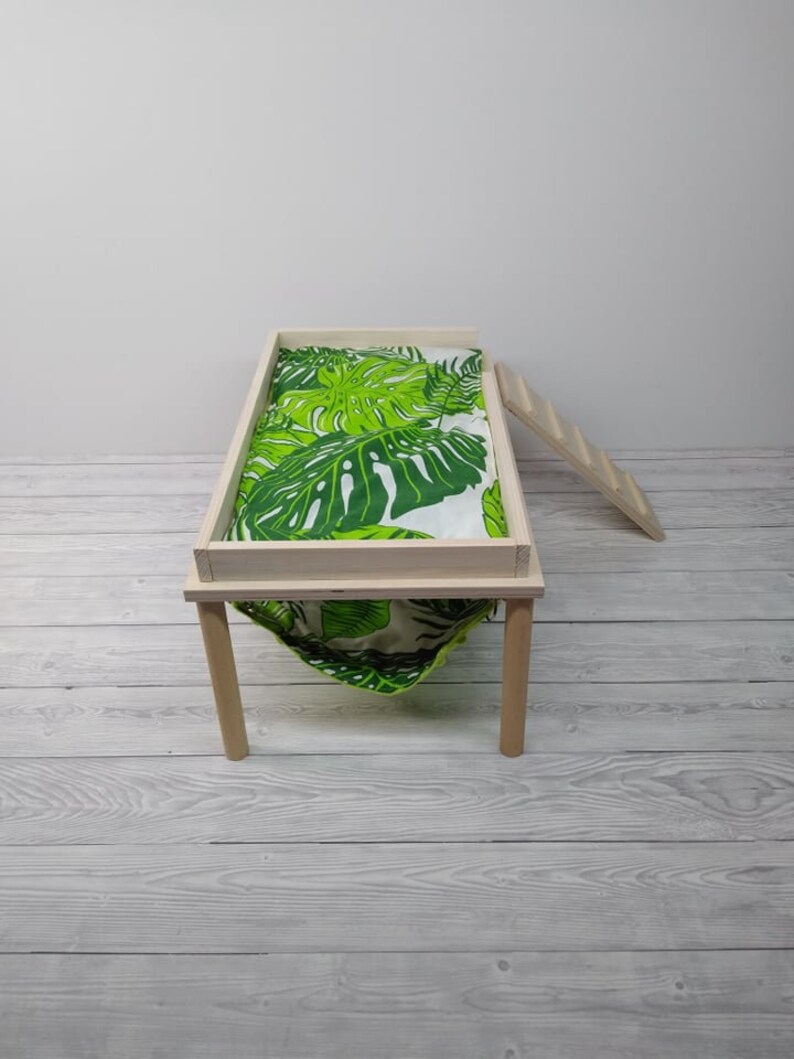May include: A small wooden pet bed with a green and white tropical leaf print fabric. The bed has a rectangular frame, four legs, and a hammock-style bottom. A small wooden ladder is leaning against the side.