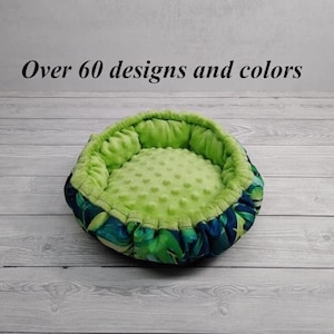 Peut inclure: Un lit pour animal de compagnie rond de couleur verte et bleue avec une doublure douce et moelleuse. Le lit a un bord surélevé. Le texte "Plus de 60 designs et couleurs" est visible sur l'image.