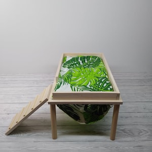 May include: A small wooden pet bed with a green and white tropical leaf print fabric. The bed has a rectangular frame, four legs, and a small wooden ladder for access. The bed is designed for small pets.