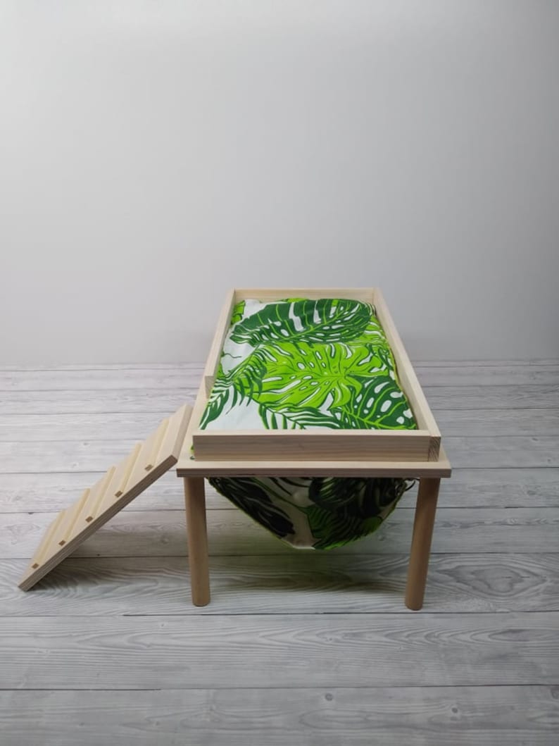 May include: A small wooden pet bed with a green and white tropical leaf print fabric. The bed has a rectangular frame, four legs, and a small wooden ladder for access. The bed is designed for small pets.