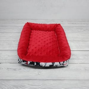 K&ouml;nnte beinhalten: Rotes und wei&szlig;es Haustierbett mit weicher, pl&uuml;schiger Auskleidung. Das Bett hat einen erh&ouml;hten Rand und einen wei&szlig;en Stoff mit einem schwarz-wei&szlig;en Muster.