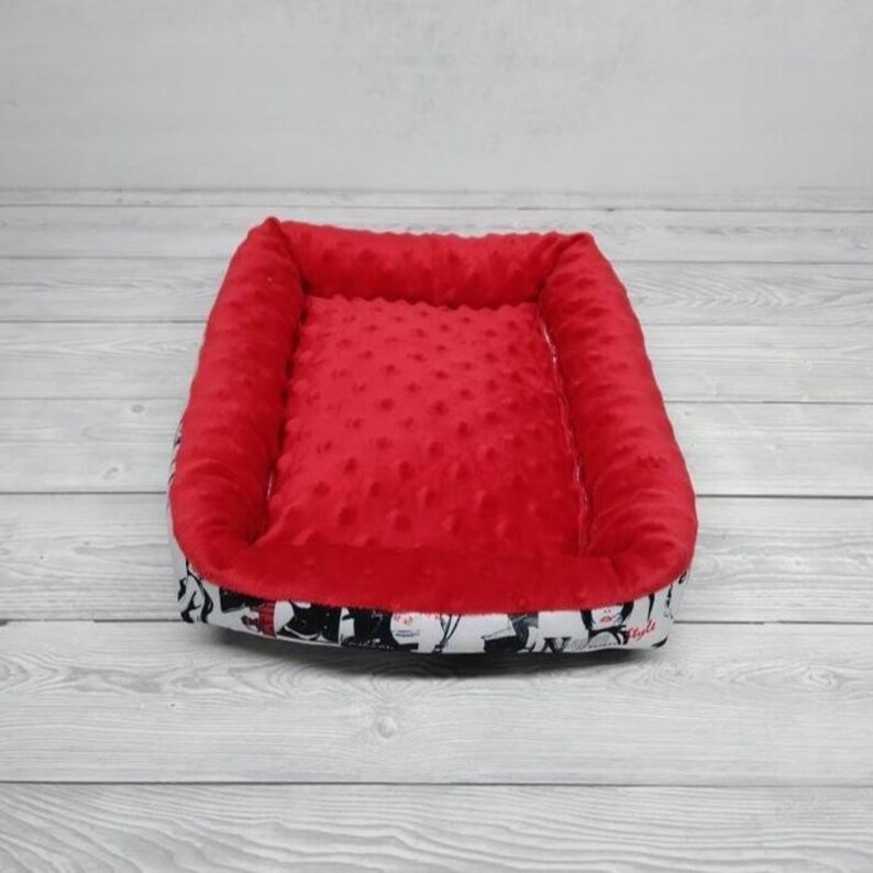 K&ouml;nnte beinhalten: Rotes und wei&szlig;es Haustierbett mit weicher, pl&uuml;schiger Auskleidung. Das Bett hat einen erh&ouml;hten Rand und einen wei&szlig;en Stoff mit einem schwarz-wei&szlig;en Muster.