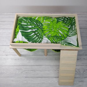 May include: A wooden pet bed with a green and white tropical leaf patterned cushion. The bed has a rectangular frame and four legs. A small wooden ladder is attached to the side of the bed.