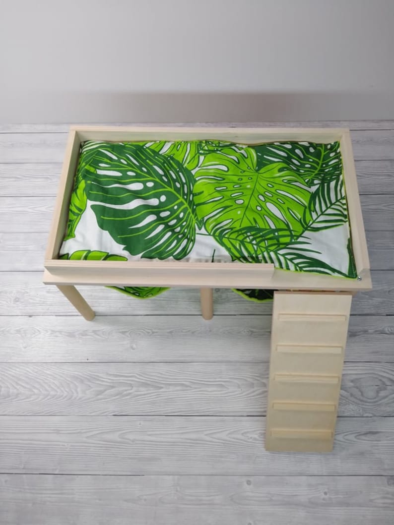 May include: A wooden pet bed with a green and white tropical leaf patterned cushion. The bed has a rectangular frame and four legs. A small wooden ladder is attached to the side of the bed.