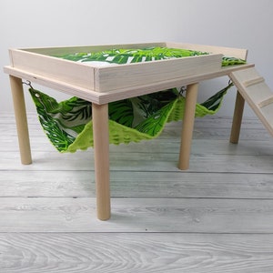 May include: A small, light-colored wooden pet habitat with a platform, hammock, and ladder. The platform and hammock feature a green and white tropical leaf pattern. The habitat has four round legs.