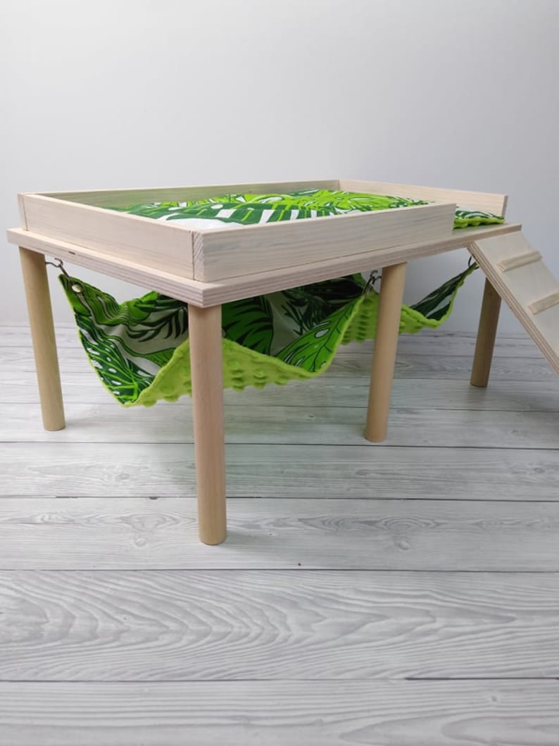 May include: A small, light-colored wooden pet habitat with a platform, hammock, and ladder. The platform and hammock feature a green and white tropical leaf pattern. The habitat has four round legs.