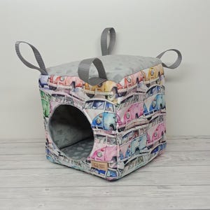 May include: A gray and white fabric pet bed shaped like a cube with a round opening. The fabric is printed with a pattern of colorful vintage Volkswagen vans.
