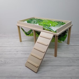 May include: A small, light-colored wooden pet habitat with a platform, ladder, and hammock. The platform has a green and white tropical leaf-patterned cushion. The ladder is angled, and the hammock hangs below the platform.