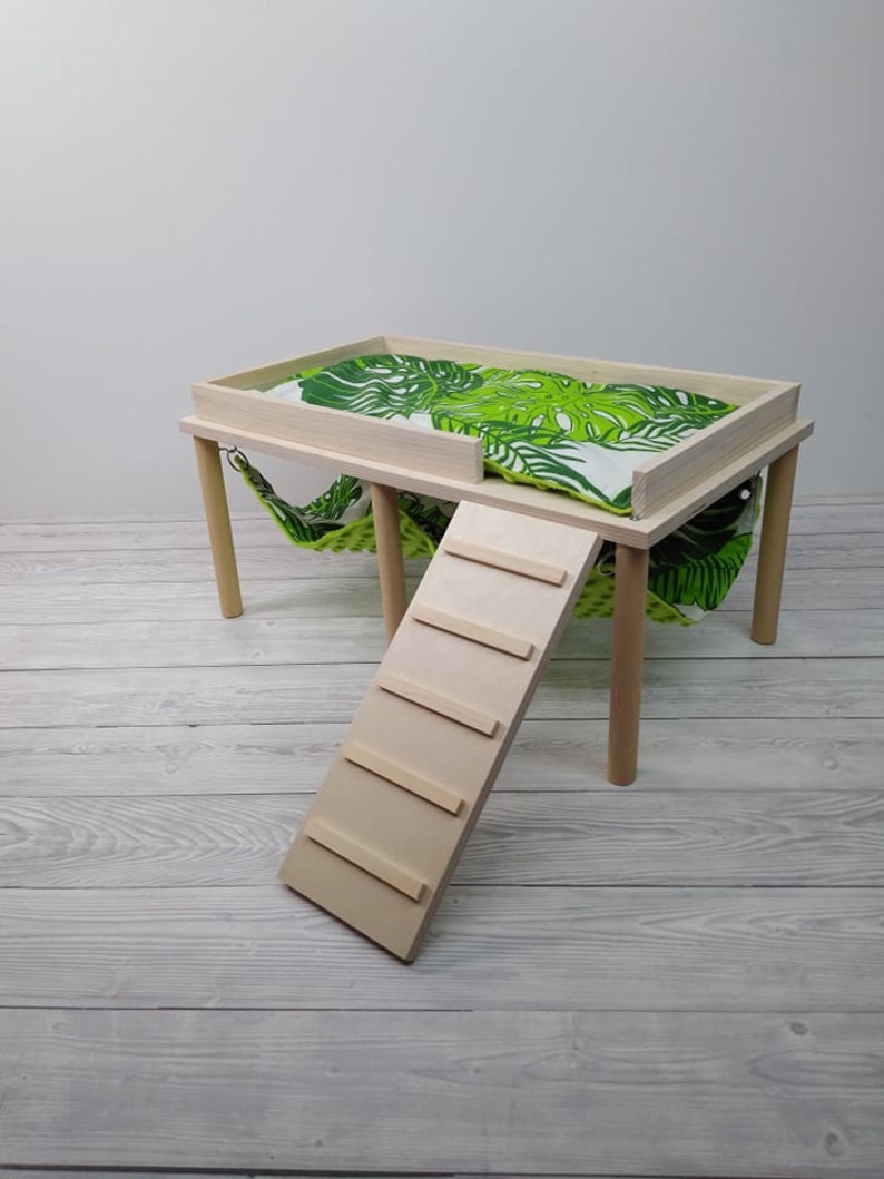 May include: A small, light-colored wooden pet habitat with a platform, ladder, and hammock. The platform has a green and white tropical leaf-patterned cushion. The ladder is angled, and the hammock hangs below the platform.