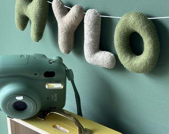 May include: A green Instax Mini 11 camera rests on a yellow and wooden box with a gold handle. Above, a felt banner spells "MYLO" in green and beige letters, strung against a green wall. The box has the word "TRAVEL" on it.