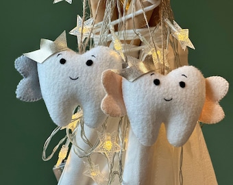 May include: Two plush, smiling tooth ornaments with wings and gold crowns. The teeth are white with black eyes and stitched smiles. One tooth has light blue wings, the other has pink wings. A string of star-shaped lights is in the background.