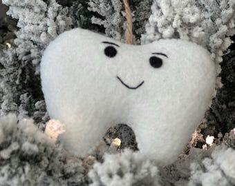 May include: A white plush tooth ornament with a smiling face, hanging on a snowy artificial Christmas tree.