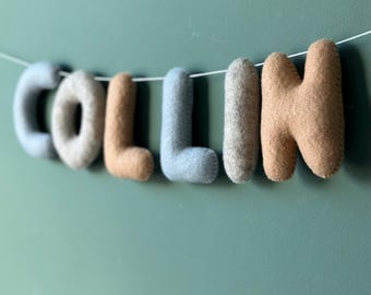 May include: A felt name banner spelling "COLLIN" in various colors, including light blue, tan, and gray. The letters are strung on a white string against a teal green wall. This decorative banner is suitable for a nursery or child's room.