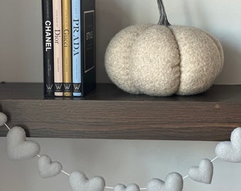 May include: A shelf with a set of four books titled "Style", "Prada", "Gucci", and "Dior" and a light gray felt pumpkin. A garland of gray felt hearts hangs below the shelf.