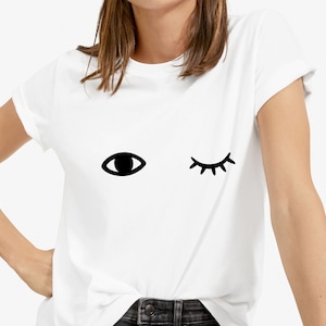 Può includere: Maglietta bianca con un occhio nero e una ciglia nera. L'occhio è aperto e la ciglia è chiusa, suggerendo un occhiolino.