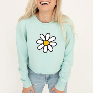 Puede incluir: Una sudadera corta de color azul claro con un gr&aacute;fico de margarita blanca. La margarita tiene un centro amarillo.