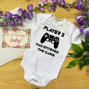 May include: A white baby bodysuit featuring the text "PLAYER 3 HAS ENTERED THE GAME" and a black game controller design. A card with the words "We're having a baby!" is also visible. The image is set against a wooden background with purple flowers.