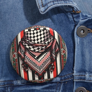 Puede incluir: Un botón redondo con un diseño detallado. Presenta un patrón de cuadros blancos y negros, rayas rojas y un diseño de bufanda con flecos. El botón tiene un borde decorativo con detalles en rojo, verde y negro. Sobre un fondo de mezclilla azul.