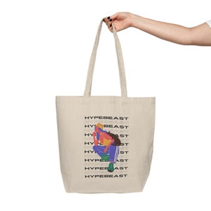 Peut inclure: Un sac fourre-tout beige avec de longues anses. Le mot "HYPEBEAST" est répété verticalement sur le côté droit. Une illustration colorée d'une personne portant un chapeau jaune, un pantalon bleu et des chaussures vertes est sur le devant.