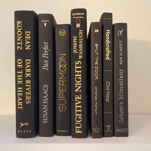 Może przedstawiać: Kolekcja czarnych książek w twardej oprawie ze złotymi napisami na grzbietach. Tytuły to "Dark Rivers of the Heart", "Supermoon", "Fugitive Nights", "Shut the Door", "Handcrafted" i "Schott's Miscellany".
