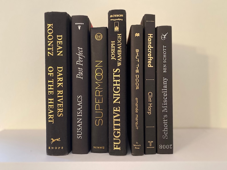 Może przedstawiać: Kolekcja czarnych książek w twardej oprawie ze złotymi napisami na grzbietach. Tytuły to "Dark Rivers of the Heart", "Supermoon", "Fugitive Nights", "Shut the Door", "Handcrafted" i "Schott's Miscellany".