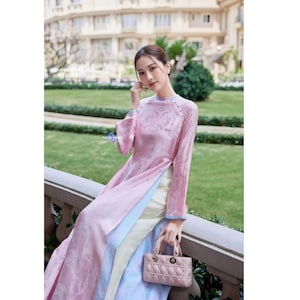 Peut inclure: Une tunique rose à manches longues avec un motif floral ton sur ton subtil, portée sur un pantalon clair. Le vêtement présente un col montant et des fentes latérales. Un petit sac à main rose rectangulaire et matelassé est tenu à la main.