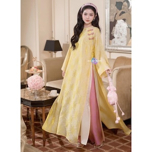May include: A long, flowing yellow dress with a floral pattern and a high slit, paired with a pink underdress. The dress features embroidered details near the neckline and the text "ALL BY HAND". A small pink purse is held in the hand.