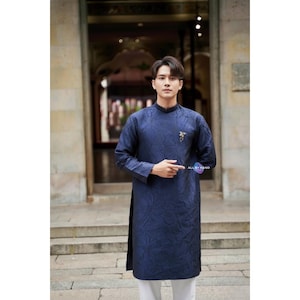 May include: A navy blue tunic with a floral pattern and long sleeves. The tunic has a mandarin collar and a decorative brooch. The text "ALL BY HAND" is visible. The tunic is paired with white pants.