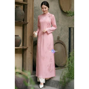 以下が含まれることがあります： マンダリンカラーと長袖の、長い薄いピンク色のドレス。 ドレスは花柄で、サイドスリットが入っています。 モデルは白いパンツと靴を履いています。 ドレスには「ALL BY HAND」の文字が見えます。