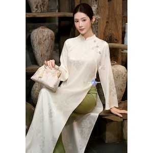 May include: A cream-coloured, long-sleeved dress with floral patterns and a high collar. The dress is paired with olive green trousers and a matching cream-coloured handbag. The text "ALL BY HAND" is visible.