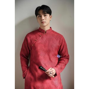 May include: A red mandarin collar shirt with an intricate floral pattern. The long-sleeved shirt features gold-colored buttons and a small pocket. The image also shows a black fan. The text "ALL BY HAND" is visible.