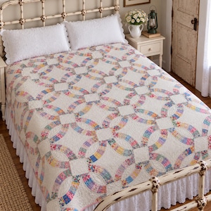 May include: A double wedding ring quilt featuring a cream-coloured background with interlocking rings composed of small, multi-coloured floral fabric patches. The quilt is draped over a bed with two white pillows and a white ruffled bed skirt.