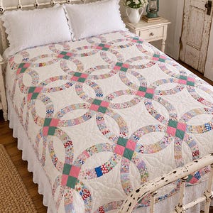 May include: A white quilt featuring a double wedding ring pattern with interlocking rings made of various small floral and geometric fabric patches in pink, blue, and green, accented by solid pink and green square corner blocks.