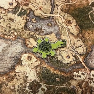 May include: A green turtle pin with a detailed shell design. The pin is on a brown and green map background.