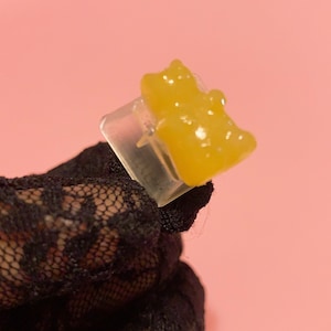 May include: A clear plastic mold with two yellow gummy bears inside. The mold is being held by a hand wearing a black lace glove.