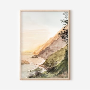 May include: A framed watercolor painting depicting a coastal landscape with a steep, grassy cliffside meeting the ocean. The scene features soft golden light, muted green and brown tones, and a silhouette of a tree on the right side.