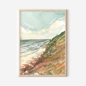 May include: A framed watercolor painting depicting a coastal landscape with a sandy beach meeting the ocean under a pale blue sky. The scene features a grassy, sloped cliffside with earthy tones of brown, green, and rust, leading down to the water.