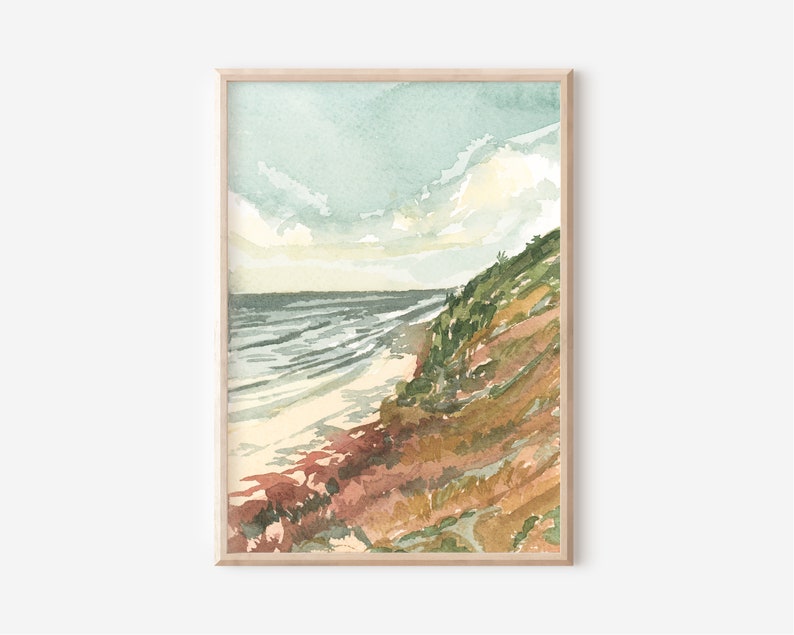 May include: A framed watercolor painting depicting a coastal landscape with a sandy beach meeting the ocean under a pale blue sky. The scene features a grassy, sloped cliffside with earthy tones of brown, green, and rust, leading down to the water.