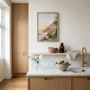 May include: A framed watercolor landscape painting of a coastal cliffside hangs above a kitchen counter. The scene features a marble countertop, a brass faucet, a beige colander with potatoes, a glass vase with a white flower, and a brown bottle.