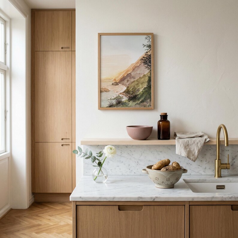 May include: A framed watercolor landscape painting of a coastal cliffside hangs above a kitchen counter. The scene features a marble countertop, a brass faucet, a beige colander with potatoes, a glass vase with a white flower, and a brown bottle.