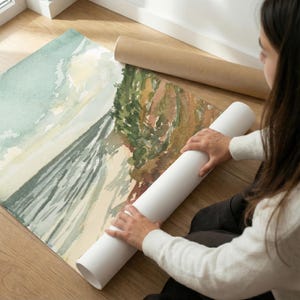 May include: A large rectangular art print featuring a watercolor landscape painting of a coastal scene with a sandy beach, dark green foliage, and a light blue sky. The print is partially rolled up, showing a smooth white paper texture on the back.