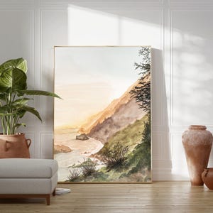May include: A vertical rectangular framed wall art print featuring a watercolor landscape of a coastal cliffside at sunset with soft orange and brown hues, green foliage, and a sandy beach meeting the ocean waves under a pale sky.