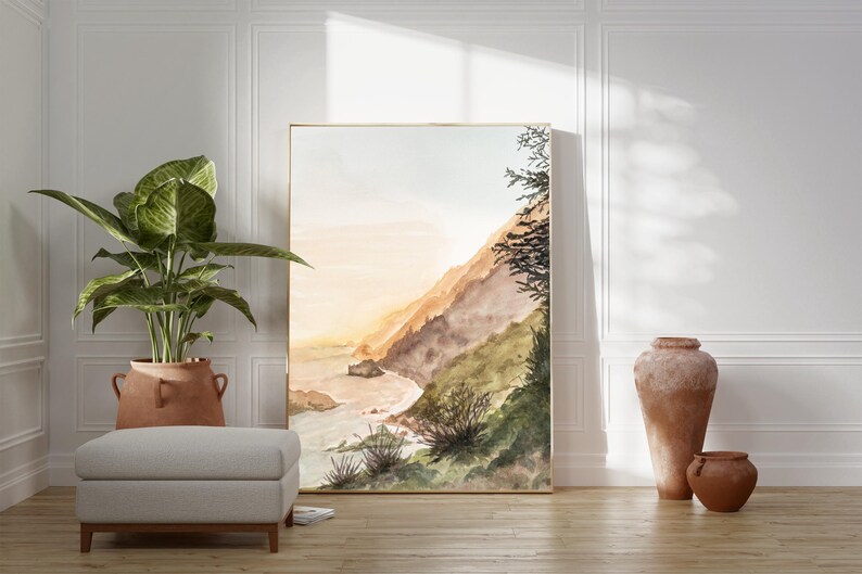 May include: A vertical rectangular framed wall art print featuring a watercolor landscape of a coastal cliffside at sunset with soft orange and brown hues, green foliage, and a sandy beach meeting the ocean waves under a pale sky.