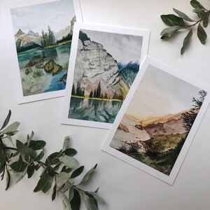 May include: Three rectangular art prints featuring watercolor landscape paintings. The first shows a mountain lake with rocks, the second a steep cliff reflecting in water, and the third a golden-hued valley with dark green foliage in the foreground.